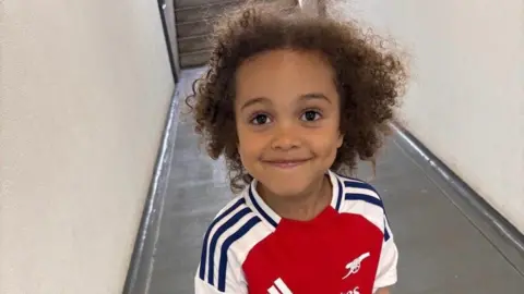 A happy smiling young boy looks into the camera. He has curly dark hair and brown eyes. He is wearing an Arsenal top.