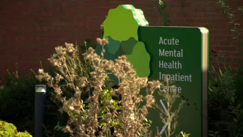 Green signage partly obscured by bush reading 'Acute Mental Health Inpatient Centre' in front of red brick building 