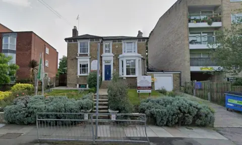 A Google screengrab of a brown brick building with large windows and a blue front door. The signage for the school is in a front garden on the grass.