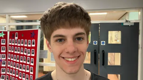 BBC Daniel is smiling. He has messy brown hair and is wearing a black top. There is a set of double doors behind him. Photos of pupils are pinned to a red board next to the doors.
