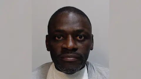 Police Scotland Momodou Bobb is staring at the camera. He is wearing a white T-shirt with a grey cotton top over it.