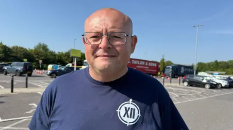 Lili Sheppard/BBC A man wearing a blue T-shirt and glasses is standing in a car park with several vehicles in parking bays.