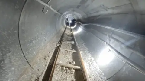 A view down an underground tunnel that is lit with bright white artificial light. There is a ladder track on the floor and various pipes and wires along the concrete walls.