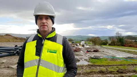 Man stands wearing hi-vis bodywarmer and a white hard hard. Directly behind him is a mucky building site and further in the background are rolling green hills. 