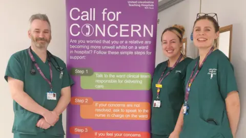 Three hospital nurses stood either side of a purple promotion poster that is advertising the call for concern service. They are each wearing green hospital uniforms with lanyards around their neck.