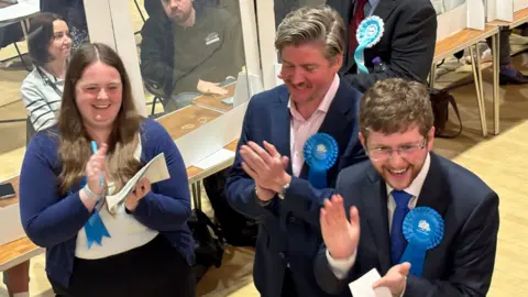 Martin Heath/BBC Two men and a woman in blue jackets with Conservative rosettes smiling and clapping at an election count with wooden tables for counting behind them.