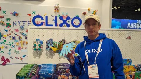 Yaron Barlev memegang burung mainan biru dan kuning, berdiri di depan dudukan clixo -nya di New York Toy Fair