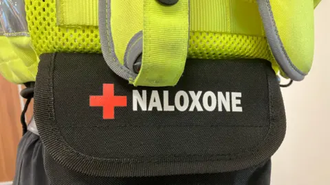 Olivia Richwald/BBC A black pouch underneath a police vest, with the text "naloxone" written in white along with a red cross. 