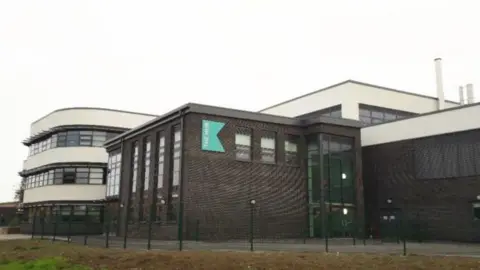 Hull City Council A school building, with brown bricks and numerous windows and a blue "Kelvin Hall" logo on the wall. In the background is a white art deco-style building.