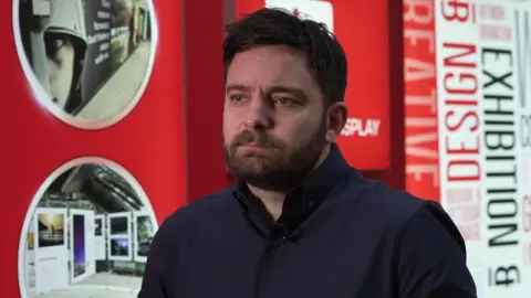 Image shows a man with a beard and moustache wearing a dark blue shirt. He is sat infront of a red background with signage 