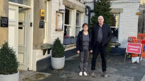 Phil Hall and his wife stand outside The Black Swan in Pickering. The building is a brick structure painted white, with two bushes on either side of the door. Mr Hall and his wife, both wearing dark clothes and glasses, stand beside a red Footpath Closed sign, which is fenced with a orange barrier.