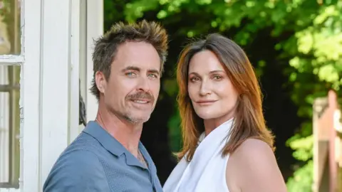 Both Jim Murray (left) and Sarah Parish are looking at the camera. Jim Murray is wearing a blue shirt and a goatee beard and short hair; Sarah Parish has shoulder length brown hair and is wearing a white top.