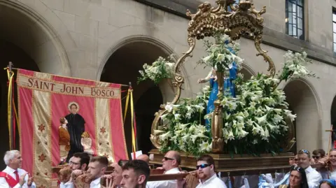 Paul Burnell/BBC Men from Manchester Italian Association carry a life-sized statue of the Blessed Virgin Mary covered with white lilies past Manchester Central Library.