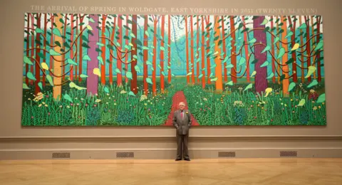 Getty Images British artist David Hockney poses in front of his painting entitled "The Arrival of Spring in Woldgate, East Yorkshire in 2011 (twenty-eleven)" at the opening of his exhibition David Hockney RA: A Bigger Picture in the Royal Academy of Arts on January 16, 2012 in London, England.