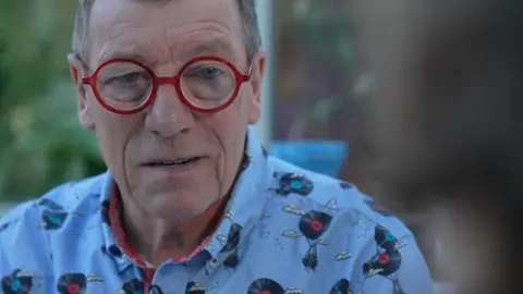 Peter Pattemore is seen wearing red round glasses and a light blue patterned shirt is pictured indoors, looking slightly off camera, with a blurred background.