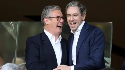 Sir Keir Starmer and Simon Harris. They are both smiling in celebration. Starmer has grey hair, wearing a dark blazer and white shirt. Harris has grey hair, wearing a navy blazer and white shirt.