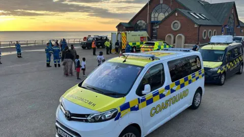 Coastguard vehicles, ambulances and rescue personnel gather outside a seafront building at sunset, with several members of the public nearby and emergency services responding to an incident on the promenade.