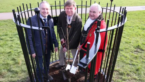 Vice Chairman of The Freemen of Newcastle Upon Tyne Nick Atkinson, Deputy Lord Lieutenant for Tyne and Wear Ed McNaught and Lord Mayor of Newcastle Henry Gallagher planting the sapling in a small circular area surrounded by a tall black fence. Nick Atkinson has short white hair and a navy blue suit and tie. He is smiling. Ed McNaught is holding a shovel. His hair is short and grey and he wears beige trousers and brown tweed jacket over a pale pink shirt. Henry Gallagher is smiling next to him. He is also holding a shovel and has short white hair. He is wearing a long red robe over a white shirt with ruffles.