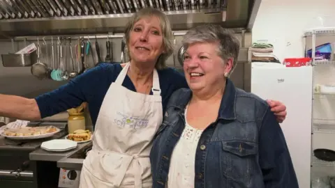 Two villagers with their arms around each other in the pub's new kitchen. There are utensils hang up and plates of sandwiches and cake in the background.