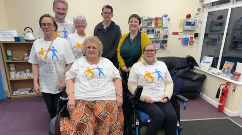 DIGS A group of campaigners and wheelchair users from the Disability Interest Group of Salisbury, with some members wearing white t-shirts with the campaign group blue and yellow logo on it. In the background there is a kettle and team cups, a sofa, a leaflet stand and a fire extinguisher next to a window. 