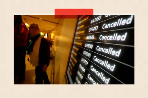 Getty Images Wide-angle view inside an airport terminal showing a large electronic departures board on the right with multiple flight listings, each marked ‘Cancelled’ in bright white text against a dark background, with flight numbers visible. To the left, several travellers walk past the display along a softly lit corridor, with luggage and coats partially visible. The perspective emphasises the repeated cancellation notices.