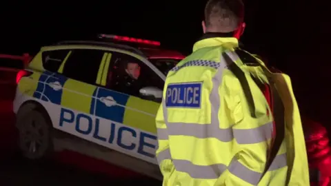 BBC A police officer in a yellow jacket looking at an officer driving a car with yellow and blue chequered stickers and the word police, in the dark, with red lights on top of the car.