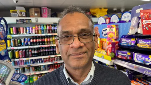 John Devine/BBC Sivanathan Kailasapillai has dark framed glasses and short dark hair, he has a grey woollen jumper and white shirt on with stock from his Post Office behind him on shelves.