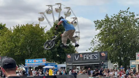 A person, wearing camouflage trousers, a blue top and a black helmet, mid-air performing a trick on his bike. There are food stalls and a big wheel in the background.
