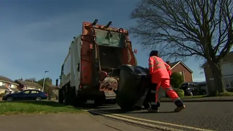 A refuse worker in high-vis overalls pushes wheelie bins towards the back of a bin lorry on a suburban street