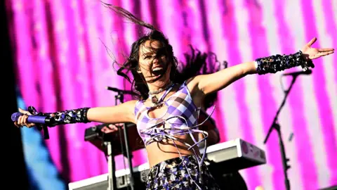 Reuters A woman in a purple crop top and purple skirt stretches out her arms in front of a pink back drop. She is holding a purple microphone in her right hand.