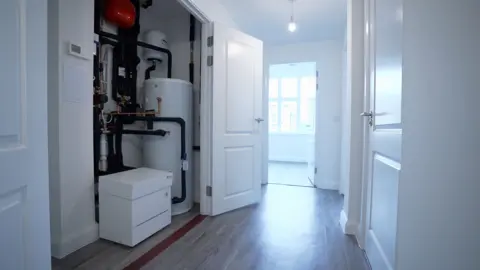 Kevin Church/BBC View from the hallway of a cupboard to the left which has a heat pump - a small white box - connected to a hot water tank via a series of pipes covered in black insulation. The system sits on a vinyl wood-like floor and is brightly lit from a window to the right