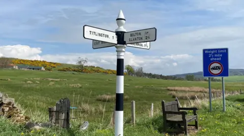 A rural landscape has a fingerpost with four directions on it in the centre of the image. The central post has alternate black and white stripes and a top that has an egg-shaped base and a pointed cone for its top half. The visible place names are Tillington (one mile), Bolton (one third) and Clanton (two and three quarters). To the right is a modern sign warning of a weight restriction ahead. There is a wooden seat between the two signs.