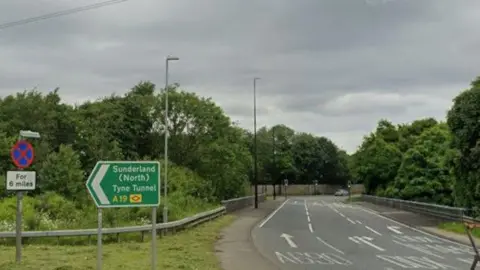 A green sign for the A19 on the A690 near Sunderland. Herrington Interchange is on the right. It is a section of a large, traffic light-controlled roundabout with trees in its centre.