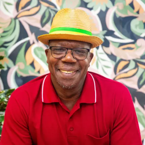 A man sits in a chair wearing a red t-shirt and a straw hat with a green ribbon. He is smiling at the camera.