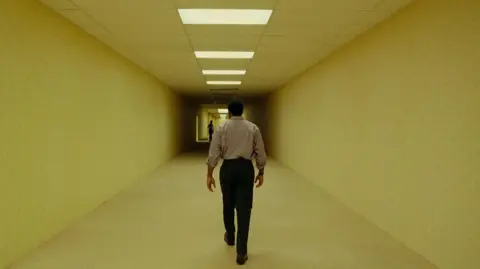 British actor Chiwetel Ejiofor as Clark, walking through the yellow hallways of the Backrooms, with a shadowy figure in the distance.