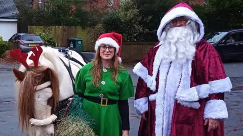 Karl Hasketh Two people stand on a residential street dressed in Christmas costumes alongside a small pony. One is dressed as an elf in a green outfit and the second is in a red Santa suit.