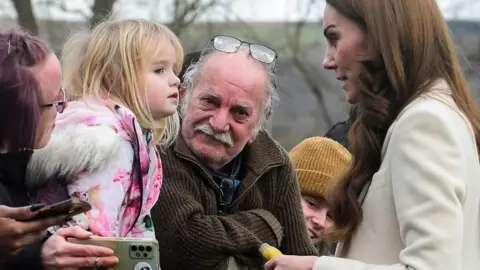 Catherine, Princess of Wales, with toy banana in hand, and watched by onlookers, chats to Lily-Rose after the unscheduled stop outside the Corgi factory in Ammanford