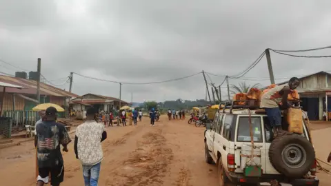 The Washing Machine Project A dirt road in the Republic of Congo. People can be seen walking in the road, and a man on a 4x4 vehicle can be seen standing on a spare tyre attached to the back of it.