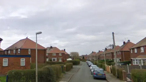 A residential street, with red brick semi-detached houses on either side, and a line of cars parked on the right-hand side of the road
