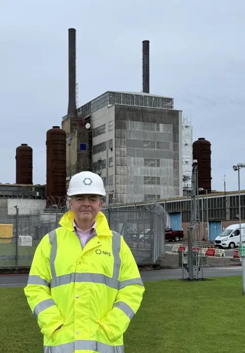 Richard Murray A man in a high visibility jacket and white hard hat stands in front of a former nuclear plant