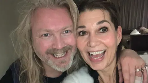 Keaton Stone A man and a woman smile at the camera with their faces close together - the photo taken is a selfie and their faces, necks and shoulders are visible. His hand is on her shoulder, with various rings and a plaster on his fingers. She is wearing a white fluffy jacket and a black necklace, while he is wearing a black t-shirt with some print on it. He has light grey hair and a beard, while she has dark brown hair.