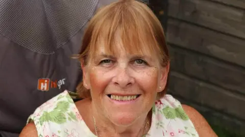 Linda Ashley-Davidson has a fringe and short hair. She is wearing a floral short sleeved dress and is sitting on a chair in front of a wooden wall.