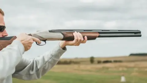 A white, brown and grey shotgun being held in the arms of someone who is preparing to fire with a finger on the trigger. The person is wearing a grey sweater. There are fields and hedges in the background.