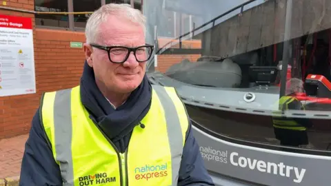 West Midlands Mayor Richard Parker wears a high vis jacket and stands in front of a National Express bus.