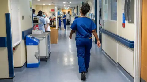 A medical professional in blue scrubs walks away from the camera along a hospital corridor. Other staff members wearing face masks can be seen to the left speaking to each other. Another medical worker is looking at a monitor in the distance.