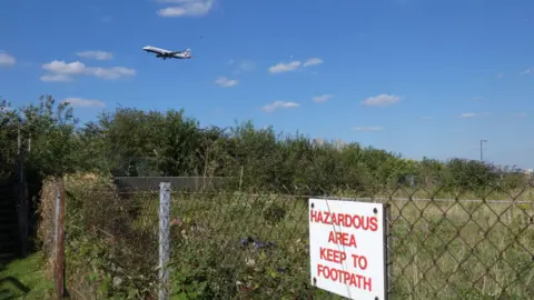 LDRS A commercial passenger plane descends through a bright blue sky dotted with small clouds, flying low above a scrubby green field bordered by a chain-link fence. A white sign with red lettering fixed to the fence reads “Hazardous area keep to footpath”. Dense bushes and overgrown grass fill the fenced-off area beneath the aircraft.