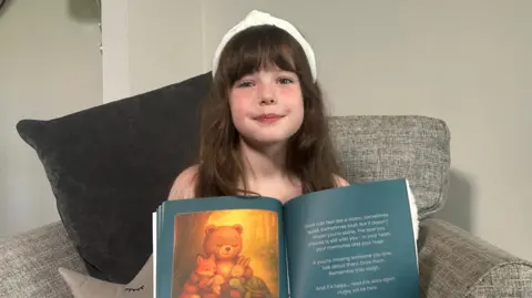 Daisy Blu Mathison holding her book open. She is sitting on a sofa and has long brown hair and is wearing a white headband. The book is open on a page displaying a cartoon bear, fox, tortoise and two rabbits all hugging.
