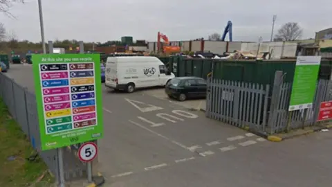 The entrance to Dorchester tip. The site is surrounded by steel fencing. There is a 5-miles-per-hour sign at the entrance. Inside the gates, several vehicles are lined up in front of a row of large dark green open-top shipping containers containing different types of rubbish and recycling.
