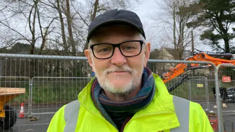 Man in high-vis jacket with glasses on. He's stood in front of construction work with an orange digger on the right hand side.