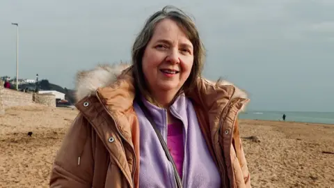 Claire Lambert, an unpaid carer, wearing a pink top, a lilac fleece and a beige coat with a fluffy collar, on the beach at Exmouth